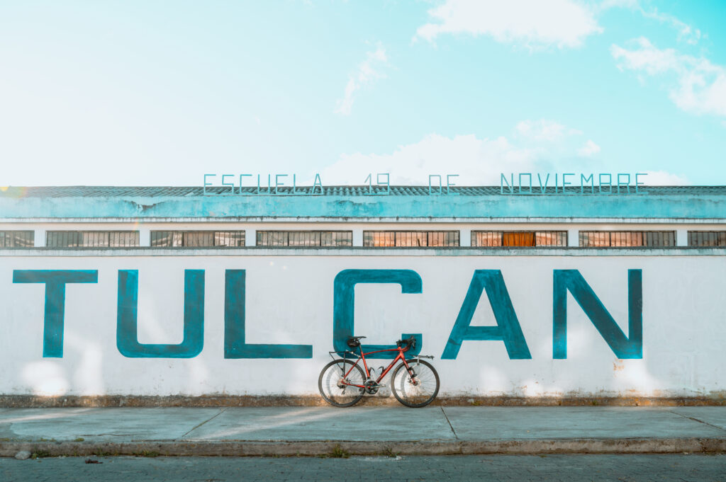 A red road bike leans against a white school wall. The word "TULCÁN" is pasted on the wall in block lettering. A metal sign on the roof reads: "Escuela 19 de Noviembre." Picture by Ben Finch