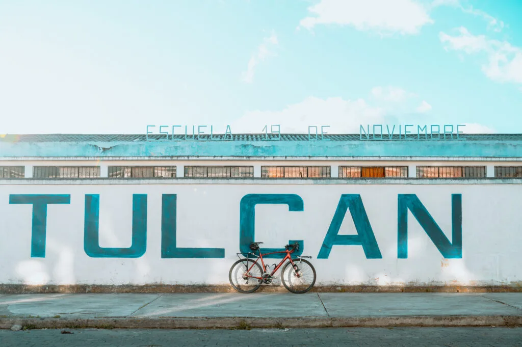 A red road bike leans against a white school wall. The word "TULCÁN" is pasted on the wall in block lettering. A metal sign on the roof reads: "Escuela 19 de Noviembre." Picture by Ben Finch Wheely Tyred Cycling laws in Ecuador