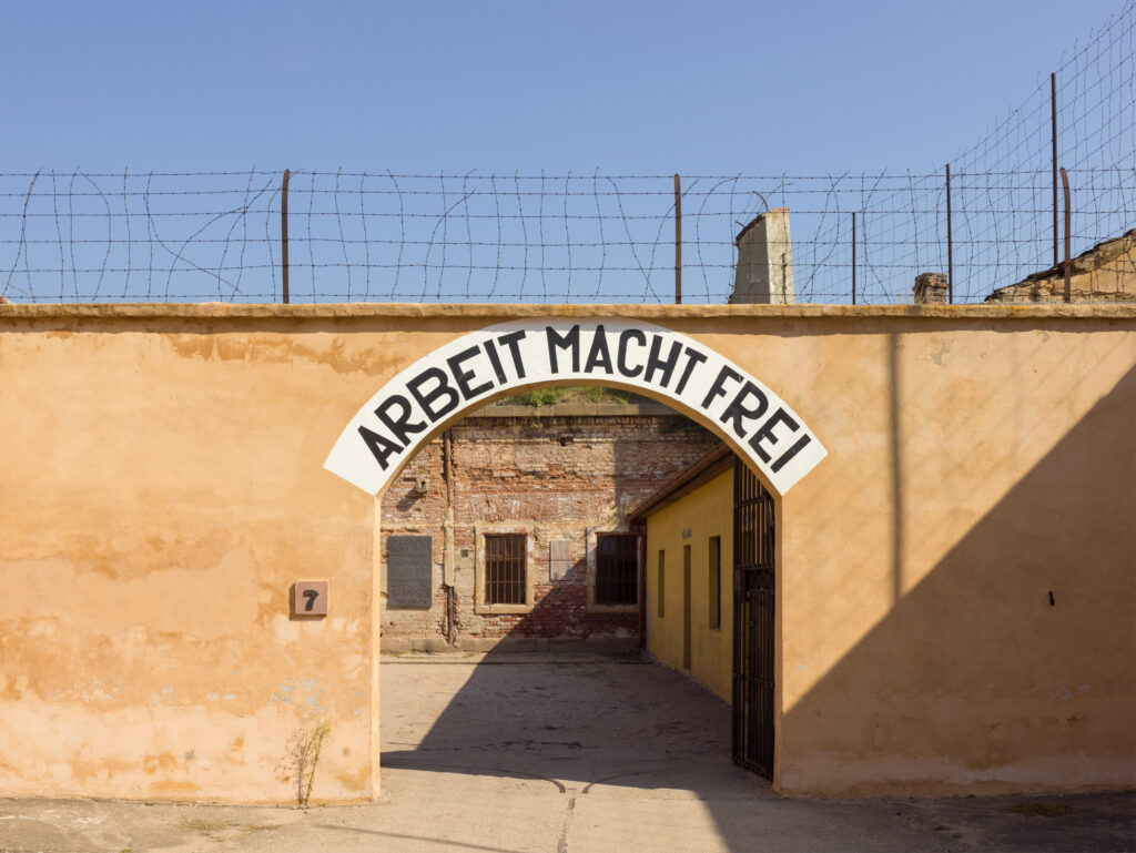 A sign above a gate in a wall reads: Arbeit Macht Frei. Barbed wire tops the wall in Terezín concentration camp
