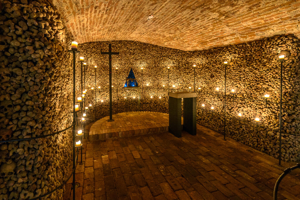 A dark room that lloks like a church, except the walls are made of skulls. The walls are lit up in the Ossuary of St James Church, Brno, and there's a cross and lecter,