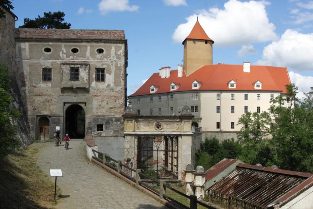 Two people on bikes ride away from one of the older parts of Veveří Castle, near Brno. The castle has a red roof.