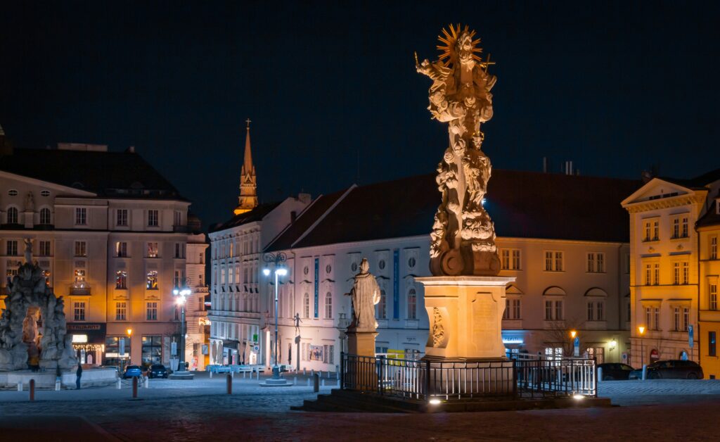 A statue rises out of a square surrounded by baroque buildings at night. Wheely Tyred