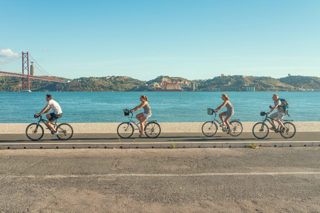 A group of people ride bikes along the seafront in Lisbon. It's a bright happy picture. Wheely Tyred cycling laws Czechia