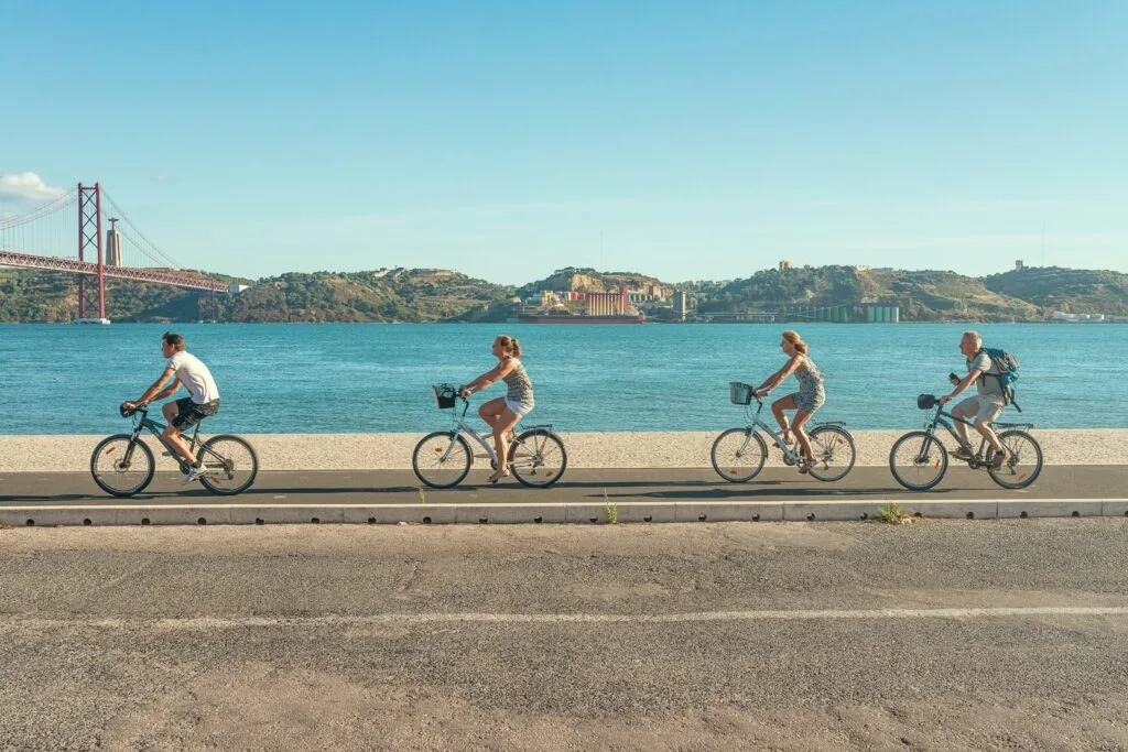 A group of people ride bikes along the seafront in Lisbon. It's a bright happy picture. Wheely Tyred bike lanes speed up emergency services