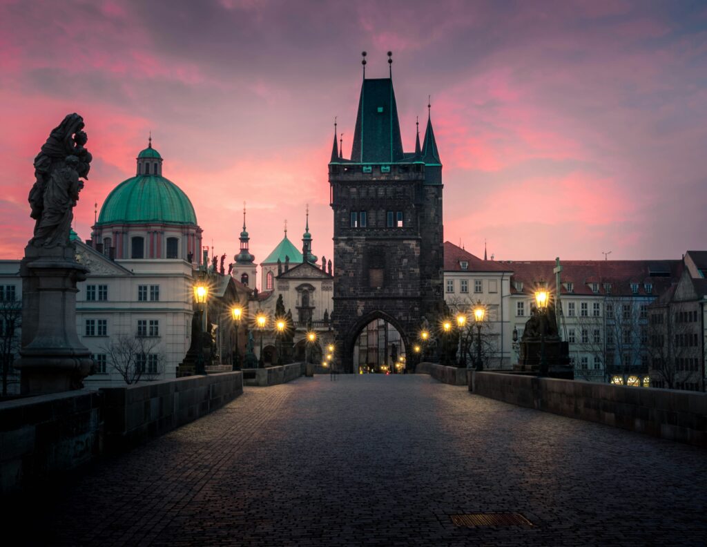 A tower at one end of Charles Bridge is lit up at dusk. Oranges and purples fill the sky behind it