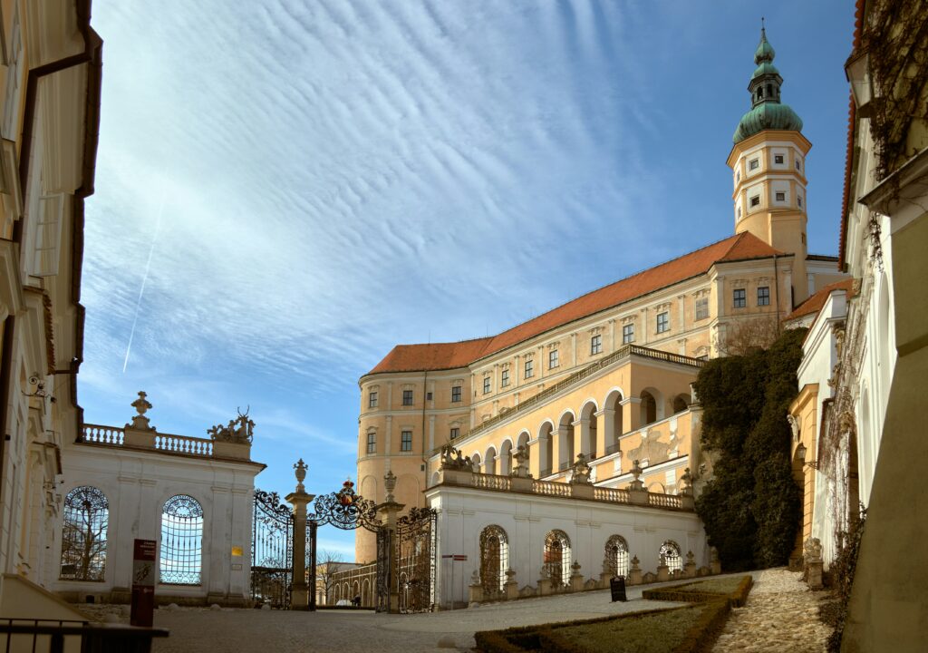 Large Baroque buildings tower over a wall and gate in a Czech town under a blue sky