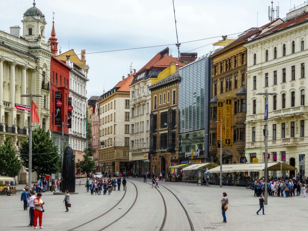 People walk across the tram tracks, past the Astronomical Clock and Baroque buildings in a large square. Wheely Tyred