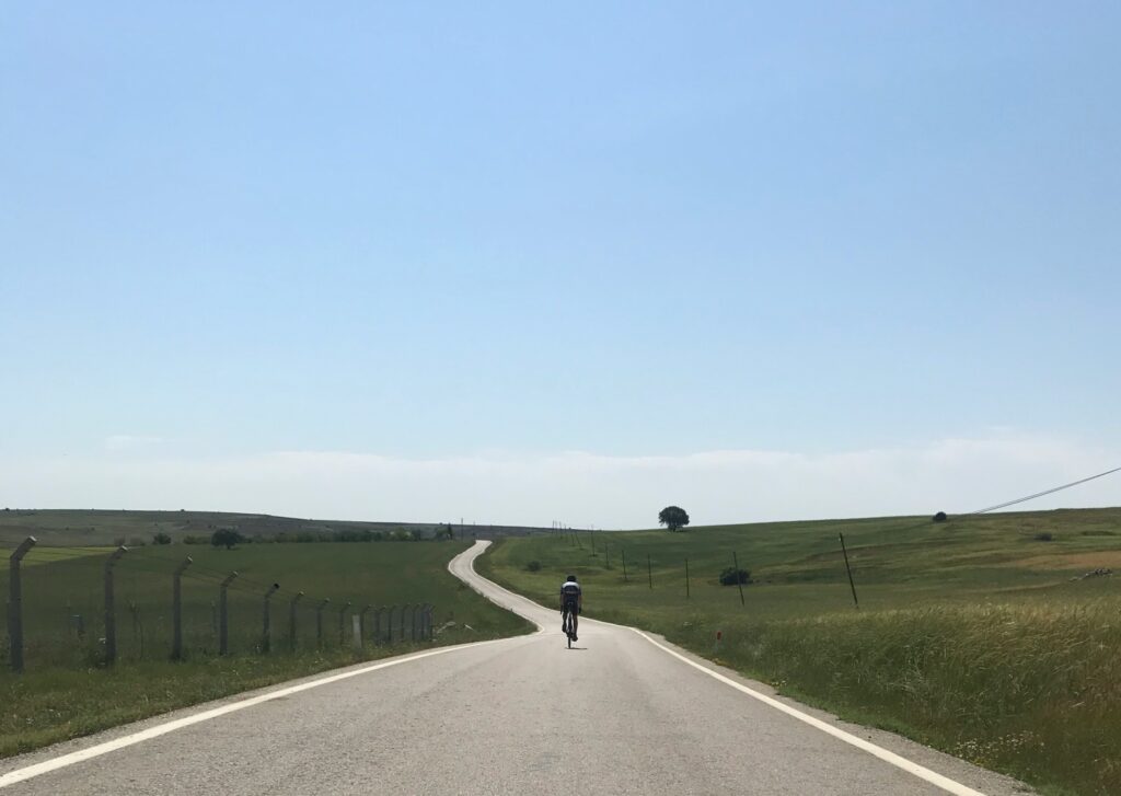 A person rides a bike along a undulating road through fields into the distance. A single tree is on the horizon. Wheely Tyred Czech cycling culture