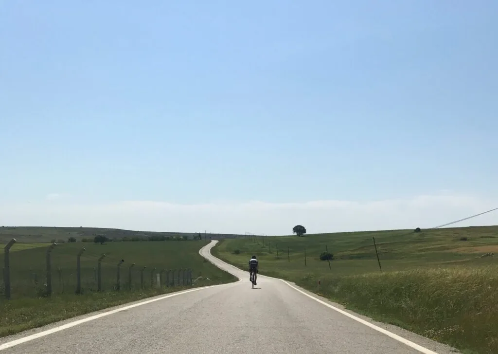 A person rides a bike along a undulating road through fields into the distance. A single tree is on the horizon.