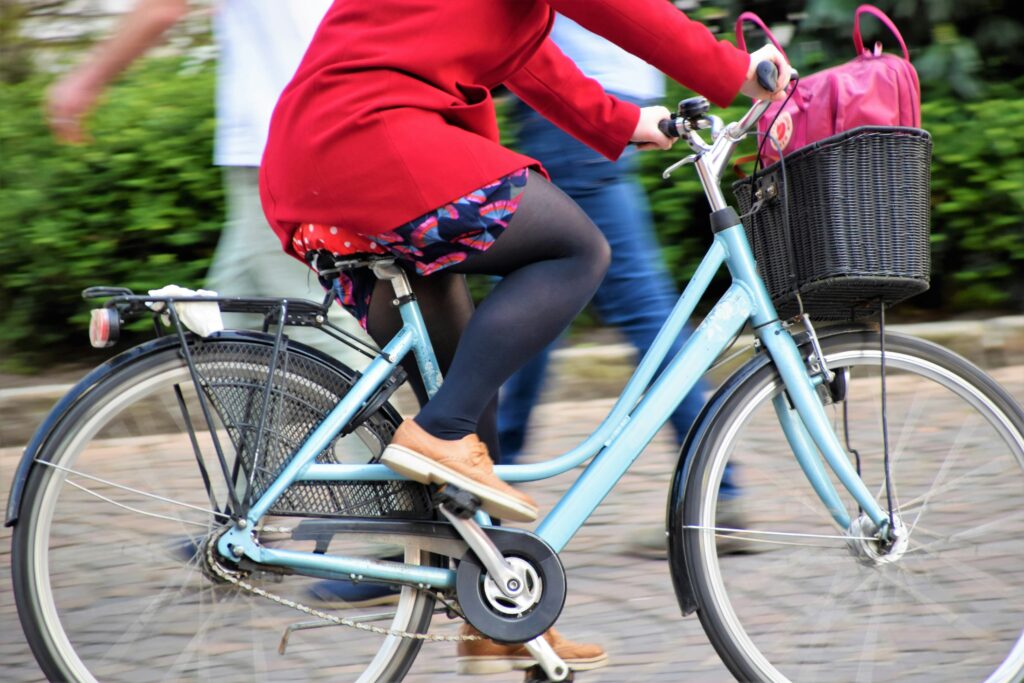 A woman ride a teal bike with a basket. She's in a red jacket and skirt.