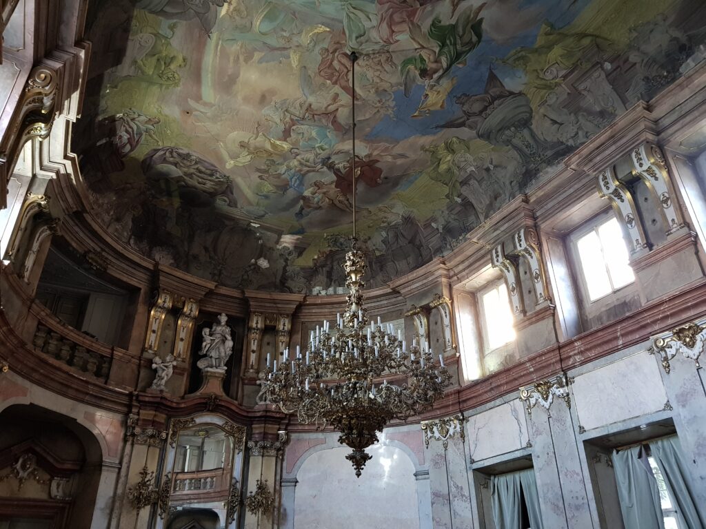Deatiled art is painted on the ceilg of the Colloredo-Mansfeldský palác in Prague. Large chandeliers hang from it.