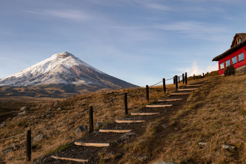 The snowcapped Volcano Cotopaxi and a red building with steps leading up to it on a clear day.