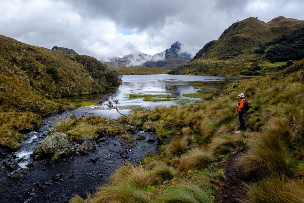 A person stands in fornt of a boggy lake, surrounde by craggy mountains, in Cajas National Park, near Cuenca, Ecuador. Wheely Tyred campsites & glamping in Cuenca