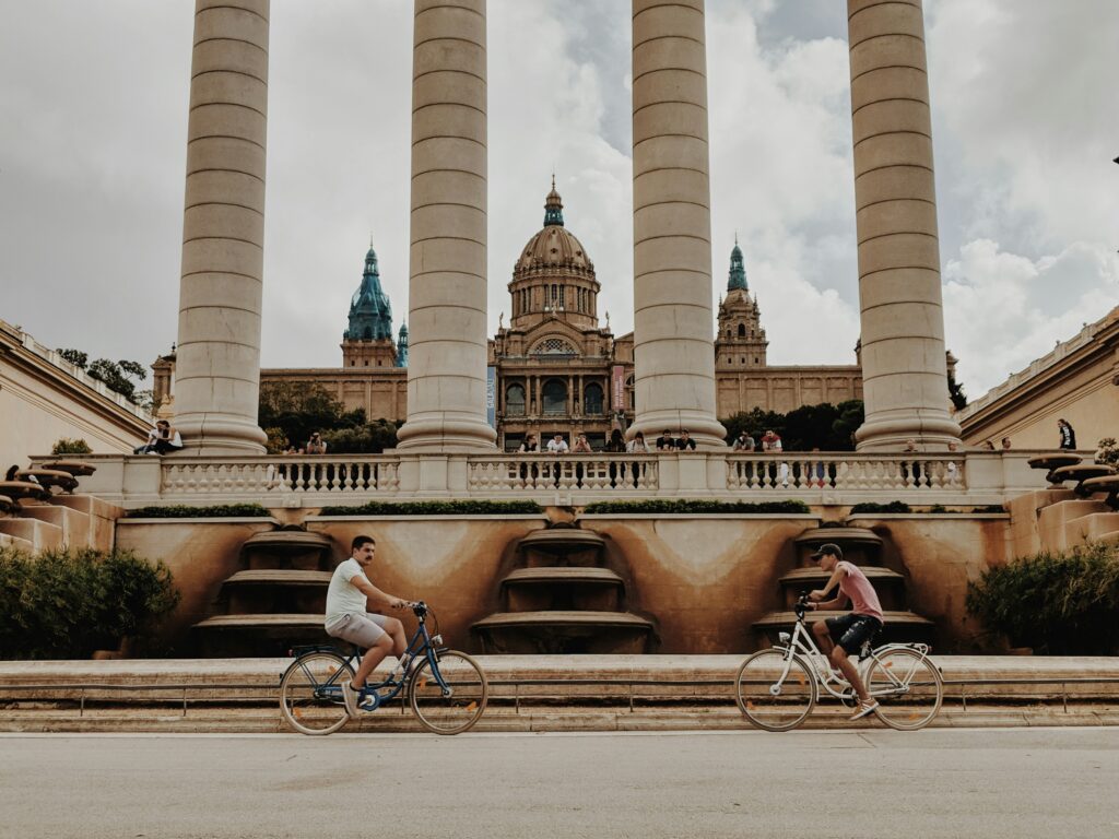 Two men ride bikes under the columns and magical buildings along Plaça Carles Buigas in Barcelona, Spain.