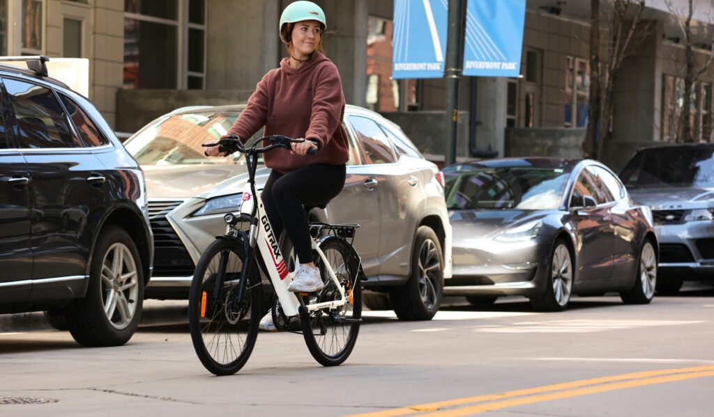 a woman rides an e-bike down the street past some parked cars. She's in normal clothes.