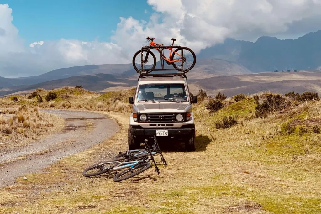 Mountain bikes lie in front of a Toyota pickup. There's on on e the roof too. The foothills of Cotopaxi surround the truck