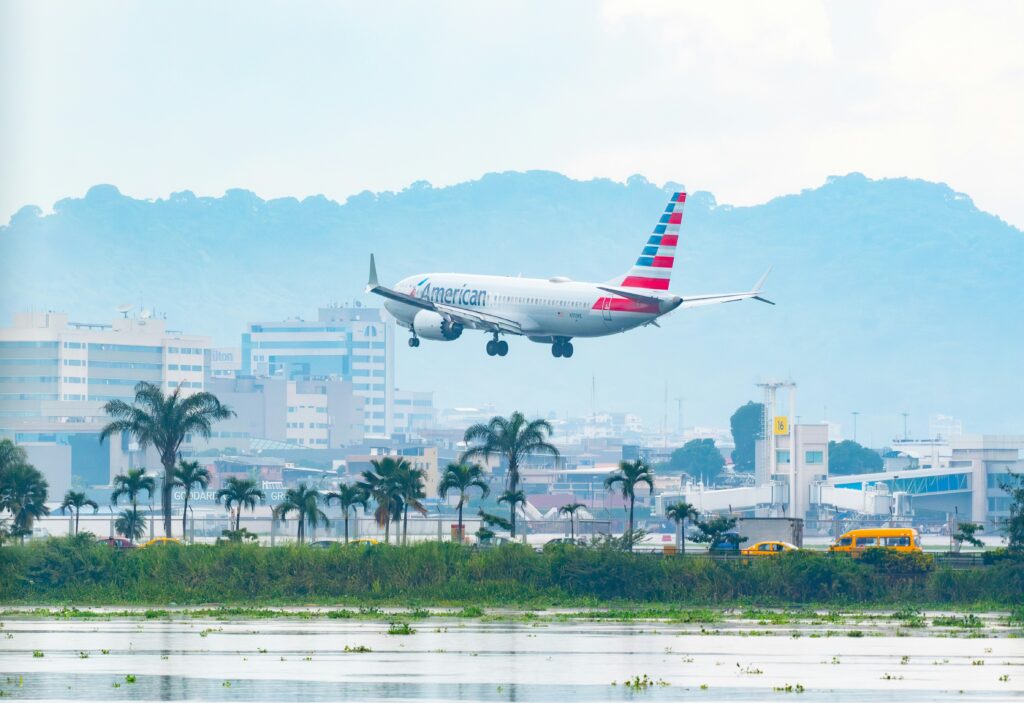 A plane lands in an airport with palm trees, the city and hills rising in the distance.