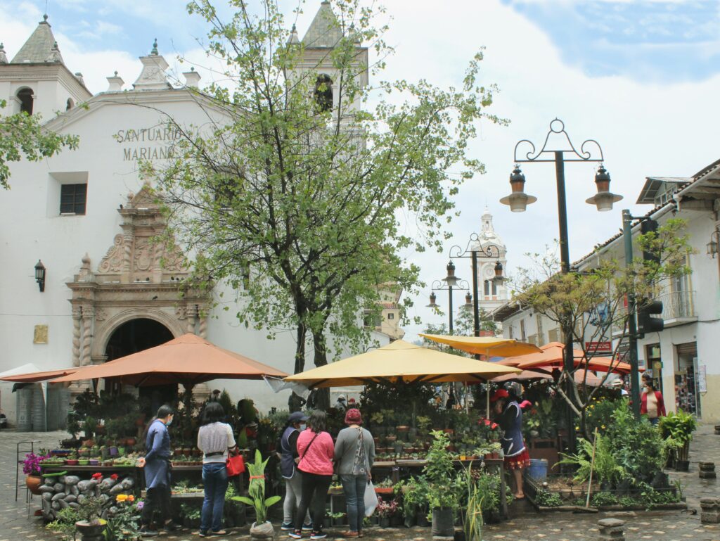People gather around stalls at the flower market under a white church in Cuenca, Ecuador.