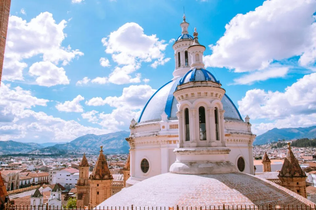 The blue and white domes of the second cathedral in Cuenca, Ecuador, are framed by a blue sky full of white fluffy clouds.