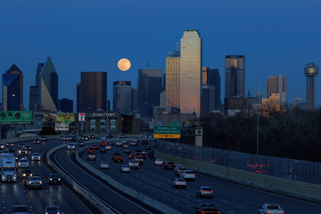 A busy highway runs into Dallas, Texas, with the moon rising over the city. Wheely Tyred Highways Destroyed America