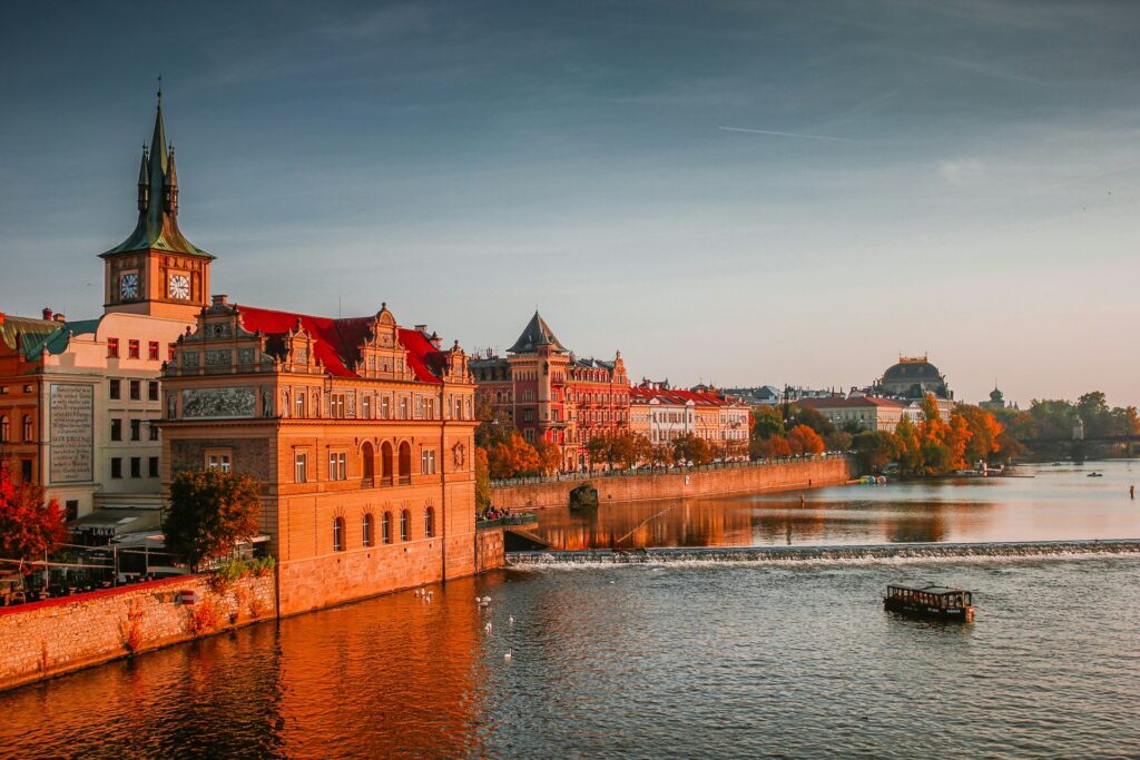 The view from the Charles Bridge in Prague at sunset. A boat floats up the River Vltava towards ancient buildings bathed in golden hour sun.