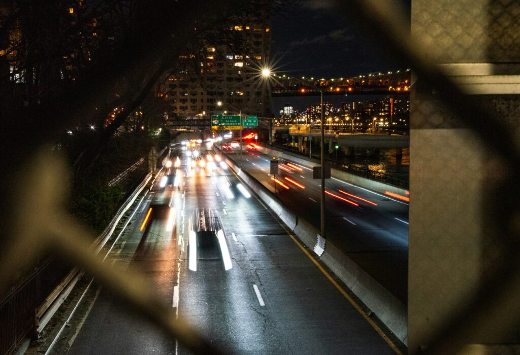 A 6-lane highway in New York at night seen through a fence. Car lights blur along it. Wheely Tyred Highways Destroyed America