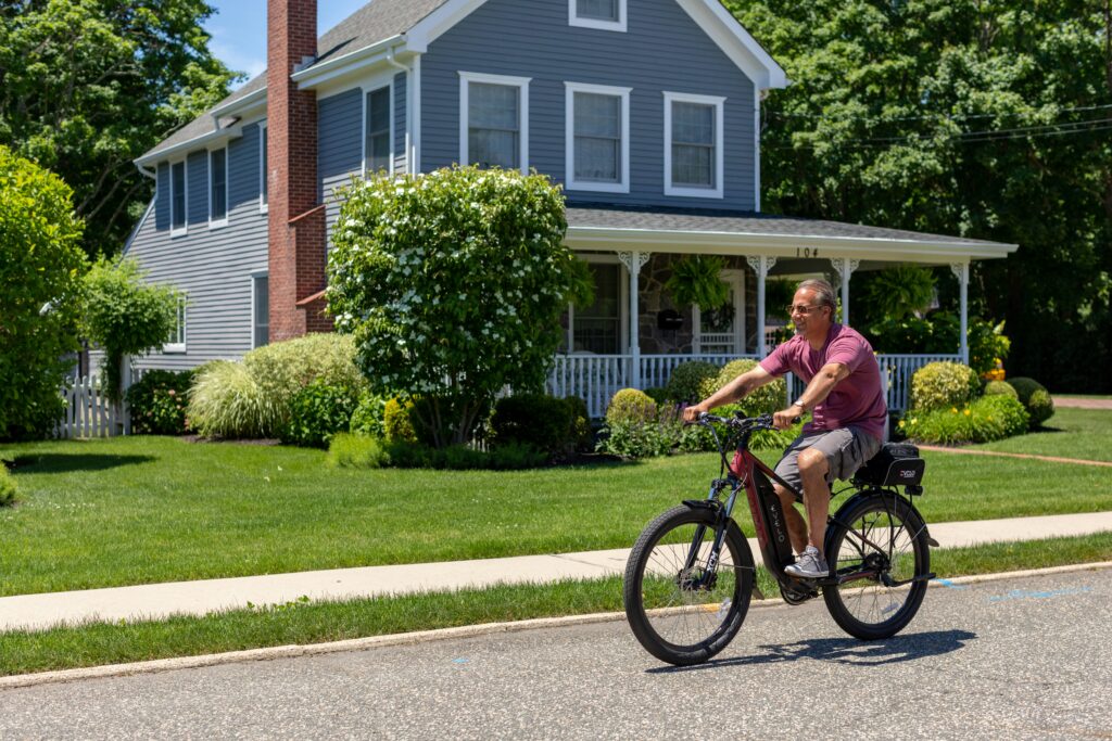 A man in shorts and a t-shirt rides an e-bike past a blue wooden house in a press shot.