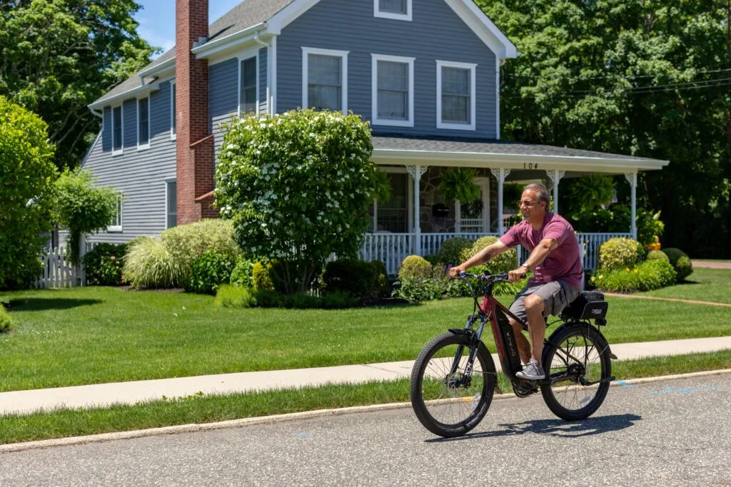 A man in shorts and a t-shirt rides an e-bike past a blue wooden house in a press shot.