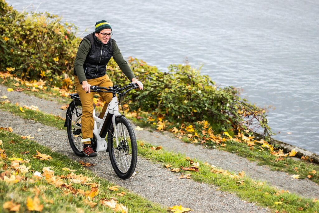 A man in normal clothes and a beanie rides an e-bike downa path lined with grass.