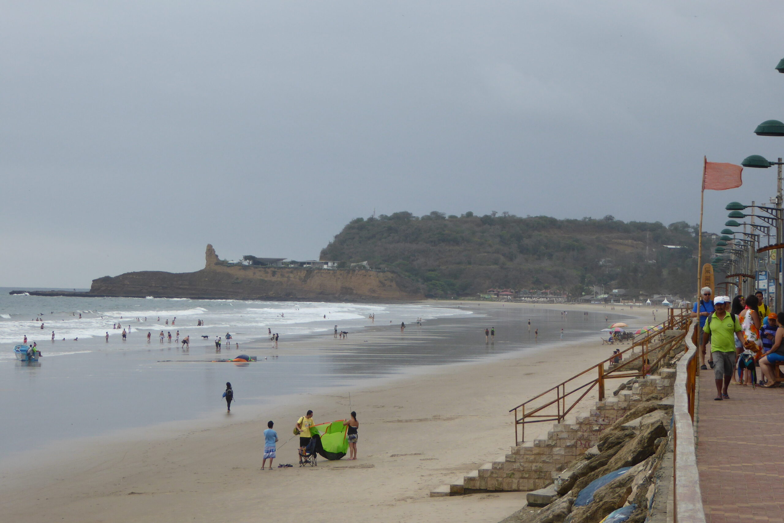 People play and walk along the beach in Montañita, Ecuador. There's a paved promenade above it.
