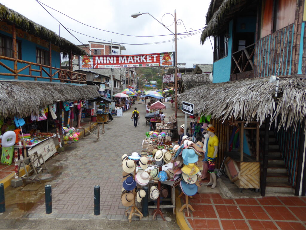 Street vendors sell hats on an empty street between blue wooden buildings with thatched roofs in Montañita, Ecuador.