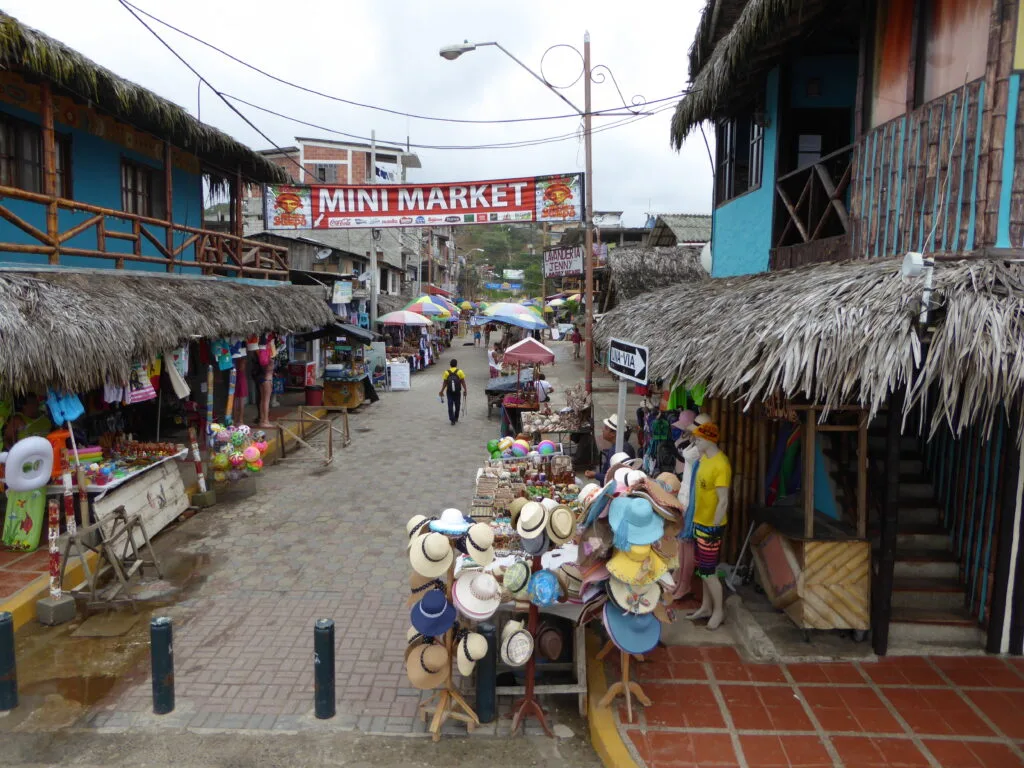 Street vendors sell hats on an empty street between blue wooden buildings with thatched roofs in Montañita, Ecuador.