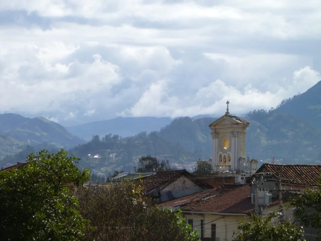 A small church tower rises in the foreground. In the distance behind it, there’s huge green valleys near Cuenca, Ecuador.