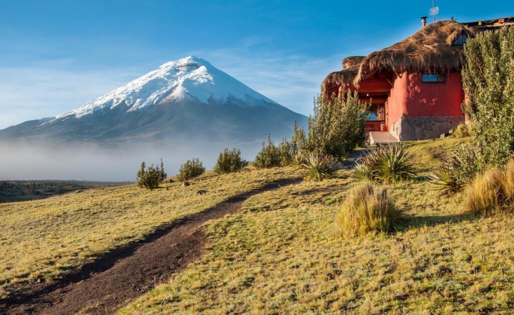 The snowcapped Volcano Cotopaxi and a red building with steps leading up to it, Hotel Tambopaxi, on a clear day.