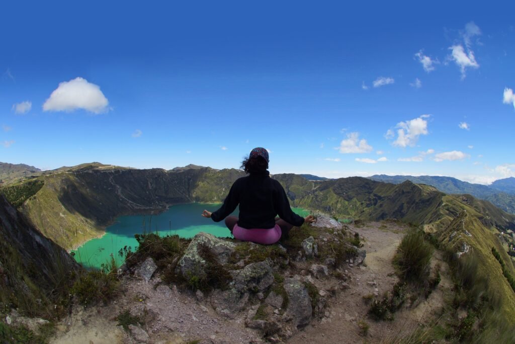 A woman sits in a yoga pose on the edge of the rim of the caldera above a bright blue lake at Quilotoa, Ecuador.