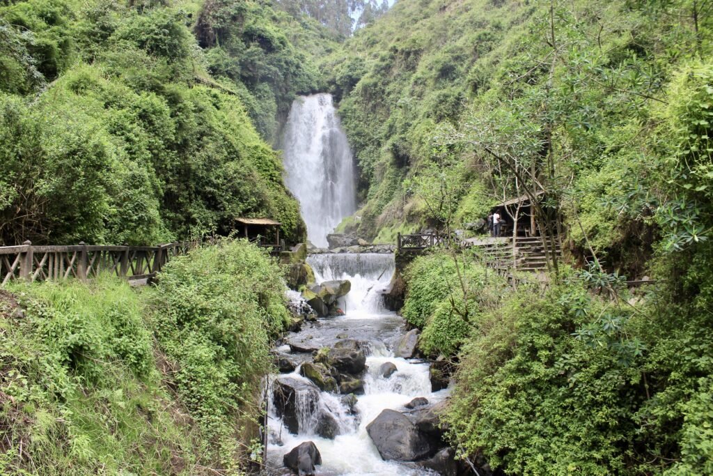 Peguche Waterfall tumbles through a forest. A bridge crosses the river after the plunge pool and and there are a few huts. It’s near Otavalo, Ecuador