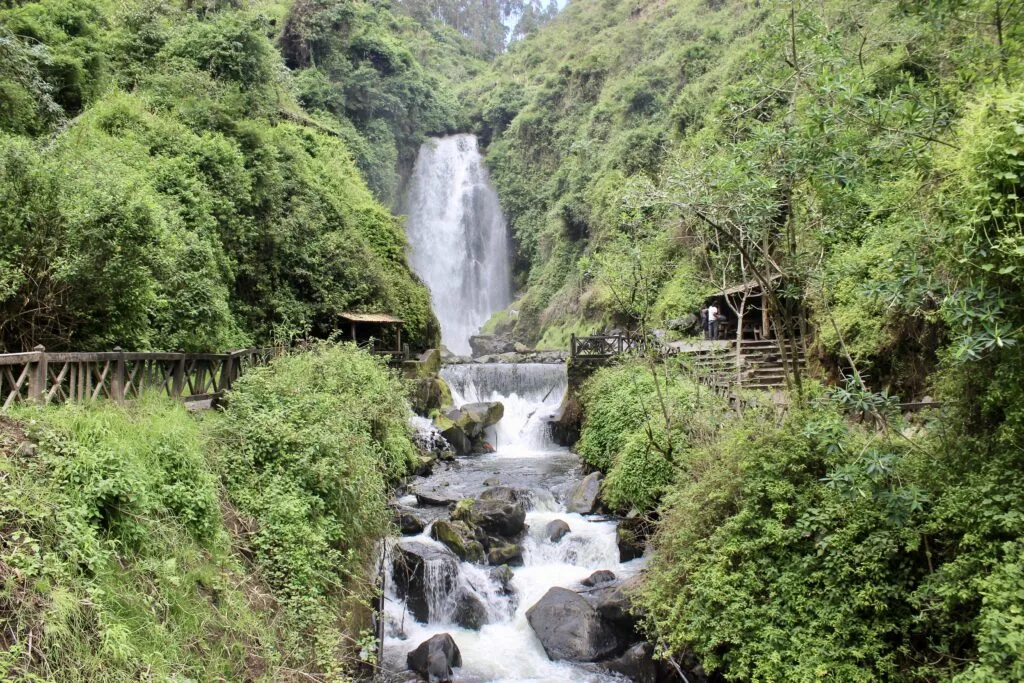 Peguche Waterfall tumbles through a forest. A bridge crosses the river after the plunge pool and and there are a few huts. It’s near Otavalo, Ecuador