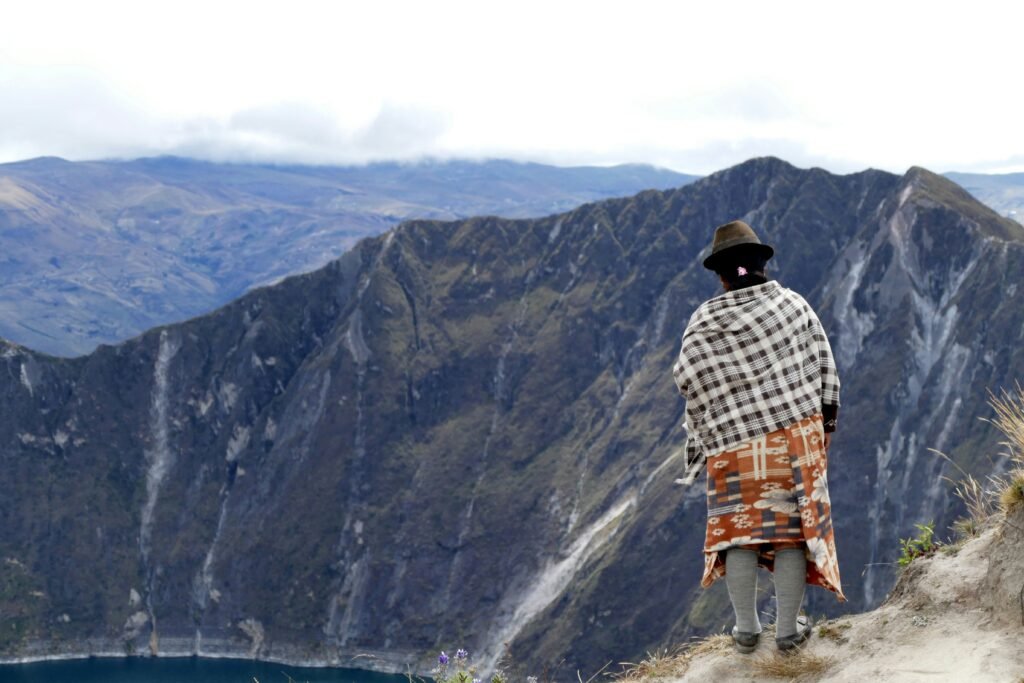 A person stands on the rim of a volcanoes caldera. They are wearing indigenous dress. A lake can be seen at the bottom of the caldera in Quilotoa, Ecuador.