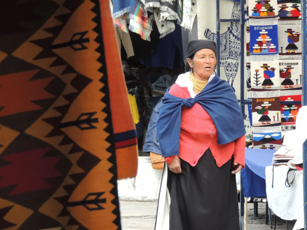 An indigenous woman shouts in a market stall covered in woven fabrics in Plaza de Ponchos, Otavalo, Ecuador