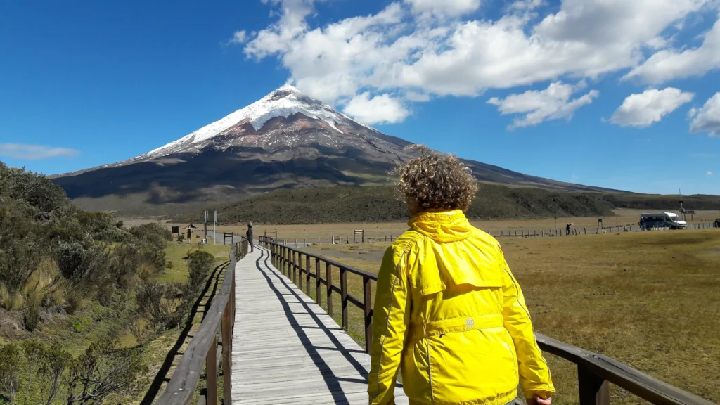 A woman in a yellow jacket walks along a boardwalk towards Volcán Cotopaxi, Ecuador