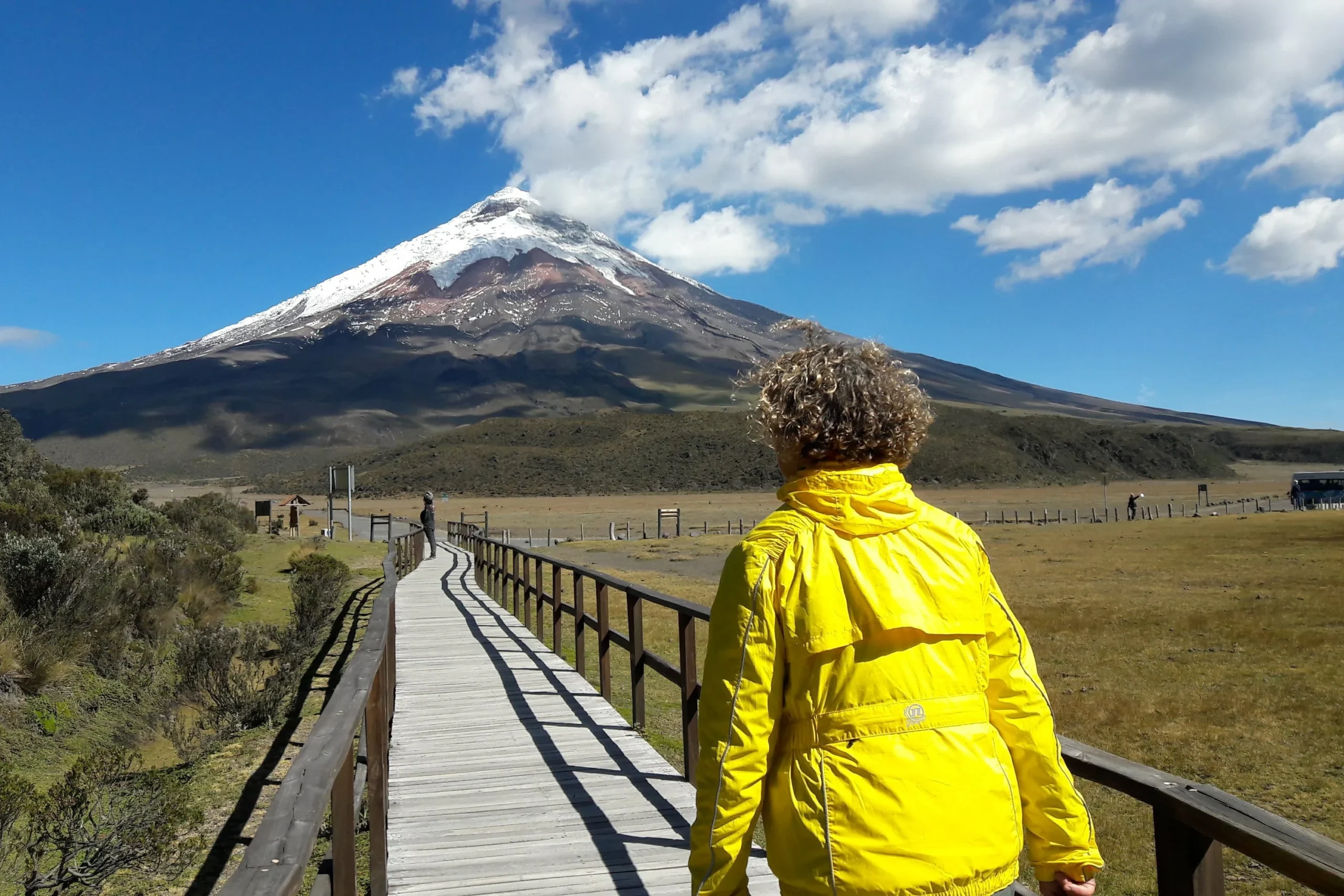 A woman in a yellow jacket walks along a boardwalk towards Volcán Cotopaxi, Ecuador