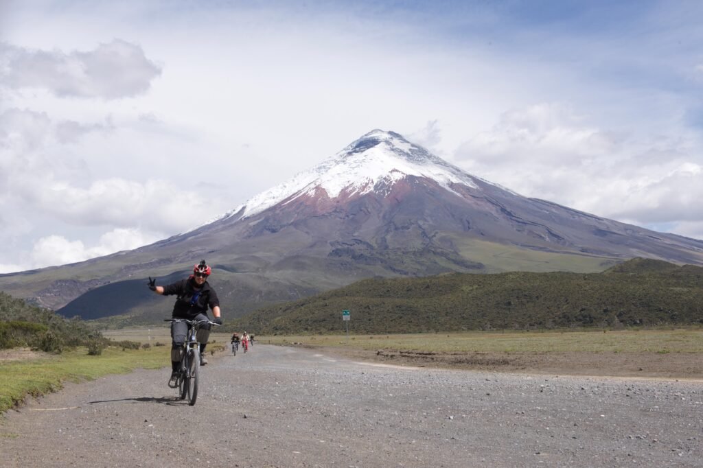 A man gives the thumbs up as he rides past a camera on a bike with Cotopaxi in the background, in Ecuador.