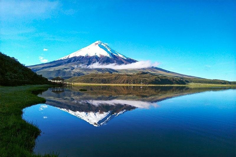 A conical, snowcapped volcano can be seen reflected in a still lake under a blue sky, in a National Park, Ecuador. Wheely Tyred Best bus tours to Cotopaxi