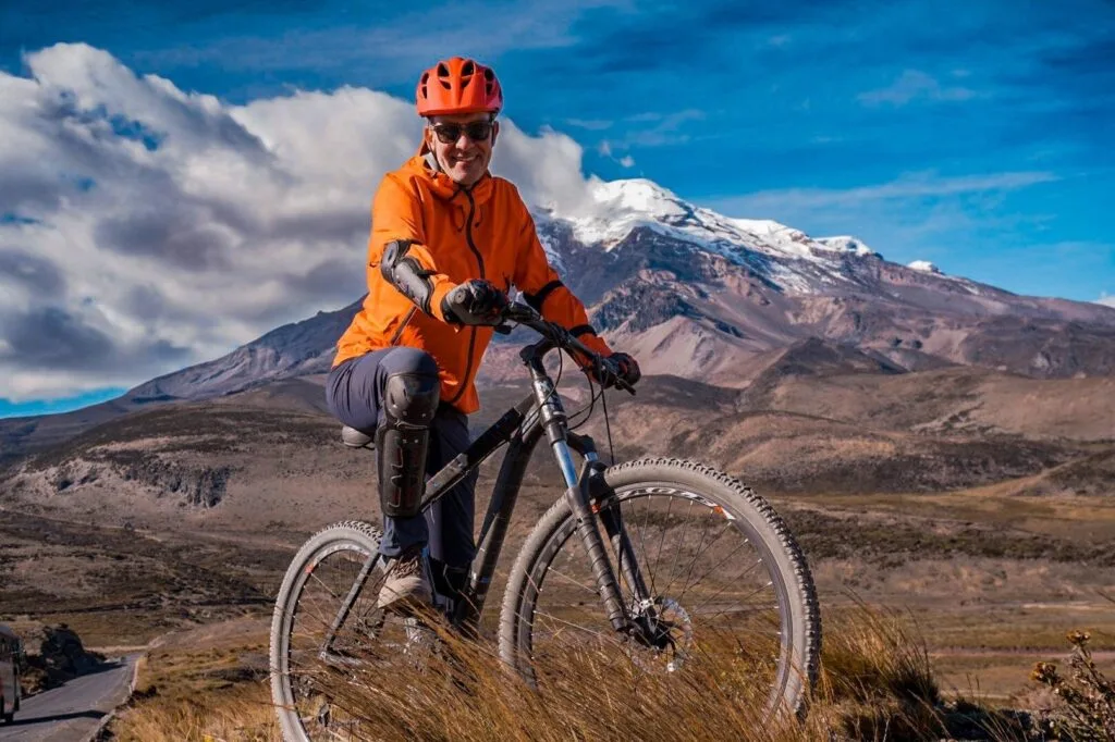 A man in orange kit sits on a mopunatin bike with clouds rolling over volcán Chimborazo behind in Ecuador. Wheely Tyred best Baños bike tours