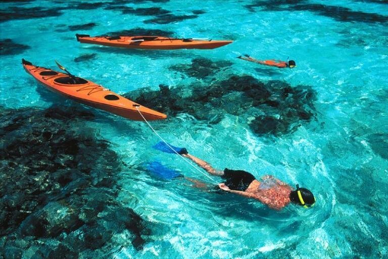 Two people snorkel in clear blue waters with their oramge kayaks attached to their wrists on Isla Isabela, Galápagos