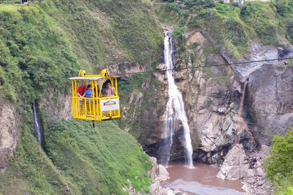 A yellow cable car travels across the Pastaza Gorge with a waterfall in the distance, near Baños, Ecuador