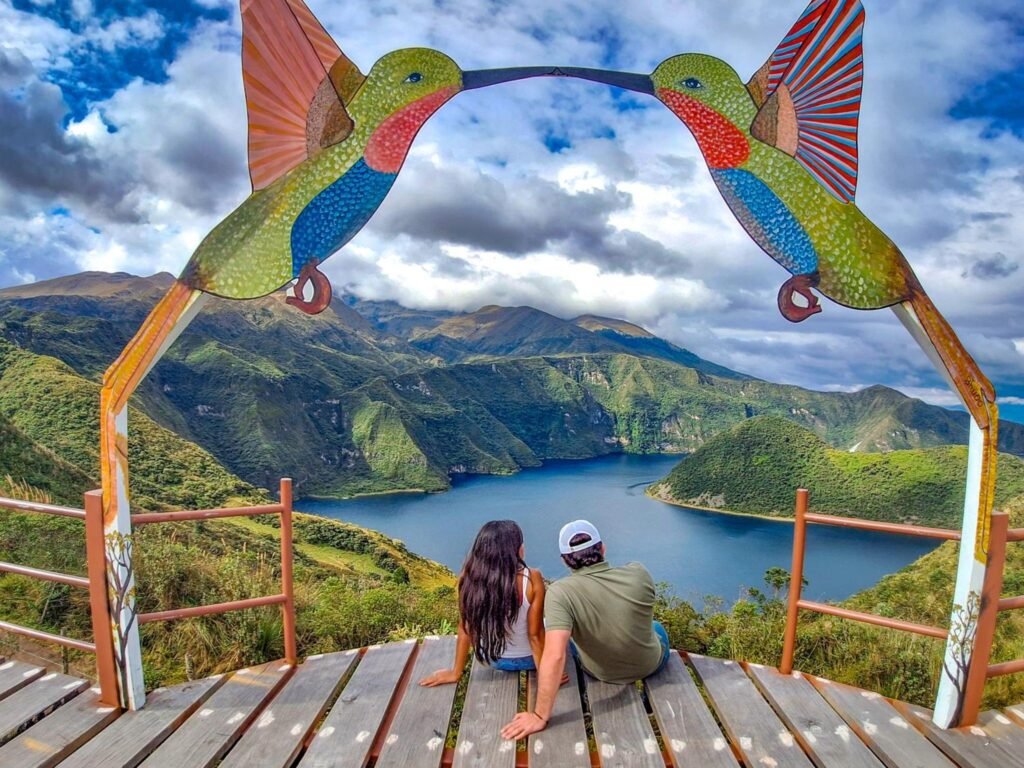 A couple sit on a wooden deck with ornmantal hummingbirds above them. A Lake is in the distance, near Cotocachi, Ecuador. Wheely Tyred stay near Laguna de Cuicocha