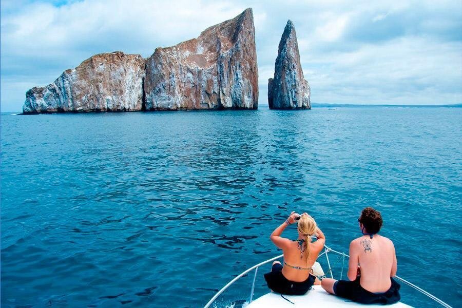 Two people sit on a boat surrounded by bright blue water looking at Kicker Rock, Leon Dormido, San Cristobal, Galapagos, Ecuador