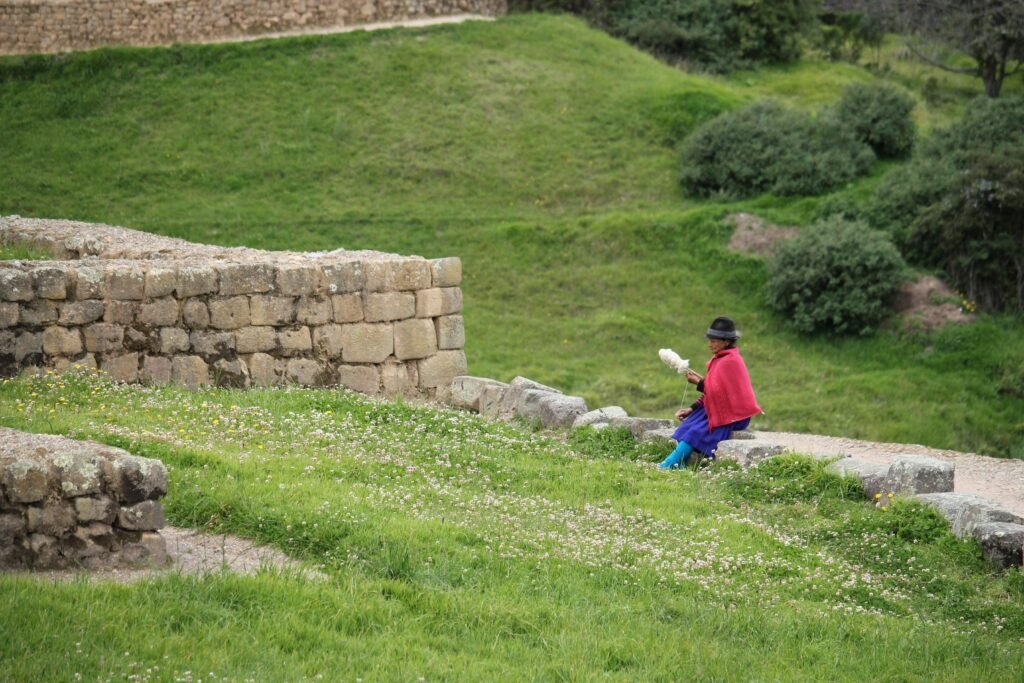 An indigenous woman in a red poncho spins some wool on the walls of Ingapirca, Ecuador